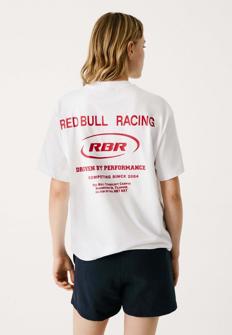 Camiseta de algodón blanca con texto gráfico rojo: "RED BULL RACING," "DRIVEN BY PERFORMANCE," y "RBR"; corte relajado y mangas cortas.