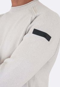 Maglione lavorato a maglia grigio chiaro con scollo rotondo, caratterizzato da un design testurizzato e una toppa in tessuto nero sulla spalla sinistra.