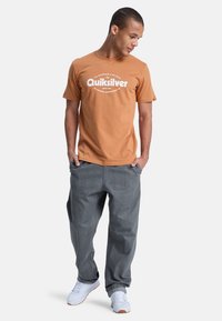 Braun Baumwoll-T-Shirt mit weißem "Quiksilver"-Logo, kombiniert mit lockeren grauen Jeanshosen und weißen Sneakern. Schlichte und lässige Kleidung.