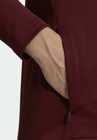Camisa burdeos de manga larga con un bolsillo lateral con cremallera; mano visible dentro del bolsillo y primer plano de los detalles del puño.
