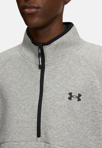 Pelēks ceturtdaļu ražots polārā rūzā ar melnu rāvējslēdzēju, tumši zilām apkaklēm un melnu Under Armour logotipu uz krūtīm. Mīksta tekstūra, sportisks dizains.
