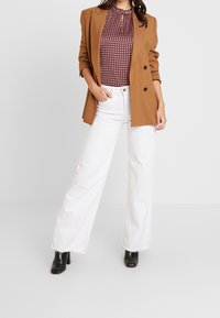 Frau in braunem Blazer, gemustertem roten Oberteil, weißen weit geschnittenen Jeans und schwarzen Knöchelstiefeln, die mit einer Hand in der Tasche steht.