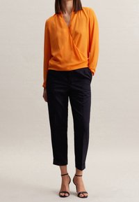 Orange wrap blouse with long sleeves, paired with tailored navy trousers and black strappy heels. Smooth fabric and tailored fit.