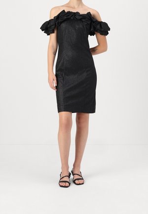 Mujer que lleva un vestido negro hasta la rodilla, con los hombros descubiertos, mangas con volantes y sandalias negras con tiras, de pie frente a un fondo liso.