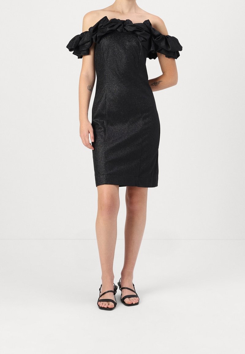 Femme portant une robe noire mi-longue, épaules dénudées, manches à volants, et des sandales noires à lanières, debout devant un fond uni.