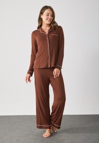 Brun, langærmet pyjamas sæt med hvid piping, der inkluderer en knap-åben toppen og løstsiddende bukser. Blødt stof, afslappet design, lomme detaljer.