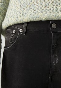 Primer plano de un bolsillo de jeans de denim negro con remaches metálicos y un suéter de punto azul claro texturizado metido dentro.