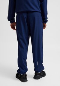 Marina atletiska byxor med en slät textur och elastisk midja. Har sidofickor och en liten logotyp på underbenet.