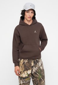 Joven con sudadera marrón y pantalones de camuflaje, con la mano en el bolsillo, también lleva una gorra gris claro, frente a un fondo blanco liso.