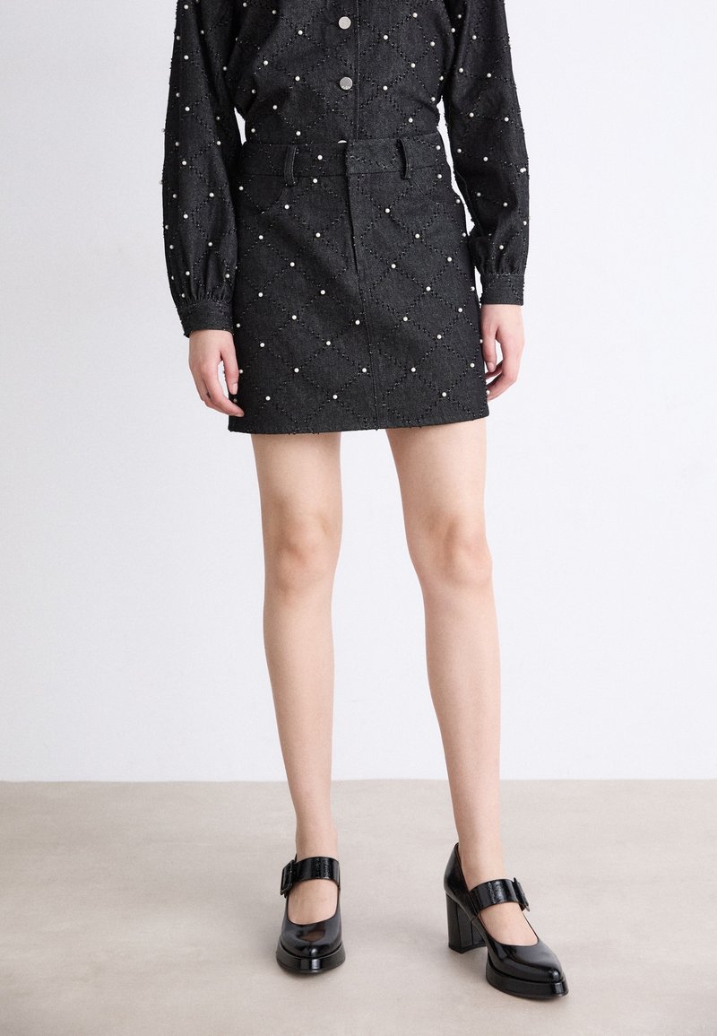 Mini-jupe en denim noir avec un motif matelassé en losanges et des accents de perles. Associée à des chaussures à talons blocs en cuir verni noir.