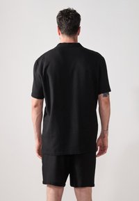 Svart t-skjorte med kort erme, avslappet passform og myk tekstur, sammen med matchende svarte shorts, vist fra baksiden.