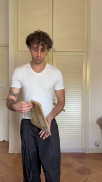 Uomo in una maglietta bianca a V e pantaloni neri, che apre una busta marrone texturizzata, con uno sfondo chiaro caratterizzato da persiane in legno.