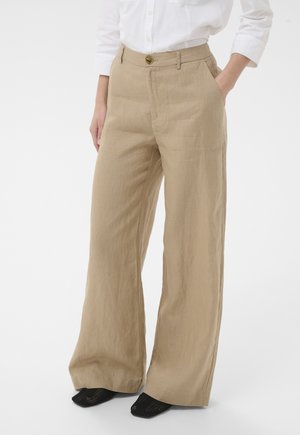 Mujer vistiendo pantalones anchos beige con bolsillos, combinados con una camisa blanca de botones y zapatos negros de tejido abierto.