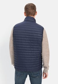 Marineblå puffervest med vandret quiltning, høj krave og en glat tekstur, båret over en beige striktrøje. Denimjeans er synlige.