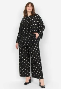 Schwarzes, plissiertes Outfit mit großen beigen Punkten, bestehend aus einem lockeren Top und weiten Hosen. Komplett mit seitlichen Taschen und schwarzen Schuhen.