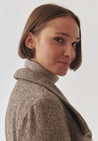 Bruine textuur coat met een grote kraag, gedragen over een lichtgrijze coltrui. Gouden ringoorbellen, kort haar, neutrale achtergrond.