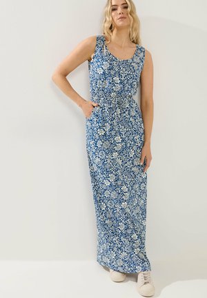 Frau trägt ein ärmelloses blaues Maxikleid mit weißem Blumenmuster, steht mit einer Hand in der Tasche und weißen Turnschuhen.