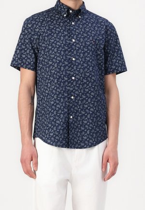 Muž v námořnicky modré košili s krátkými rukávy na knoflíky s bílým paisley vzorem a bílých kalhotách na jednobarevném pozadí.