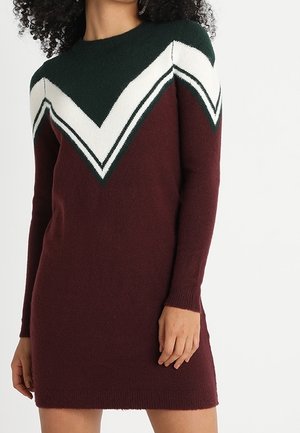 Vrouw die een lange mouw gebreide jurk draagt met een groen, wit en bordeauxrood zigzagpatroon, staand tegen een effen witte achtergrond.