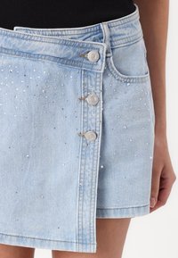 Vaalea sininen farkku mini-hame, jossa on metalliset nappikoristeet ja skarasta rhinestone-koristeita. Hameessa on klassinen viisi-taskuinen muotoilu.