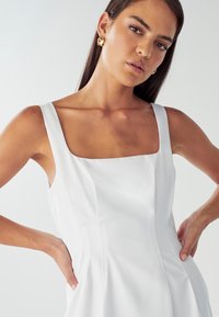 Biele bezrukávové šaty so štvorcovým výstrihom, priliehavý strih, vertikálne švy a vrecká. Model má na sebe zlaté náušnice; hladká textúra látky.