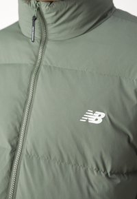 Närbild av en olivgrön puffjacka med dragkedja framtill och vit New Balance-logotyp på bröstet.