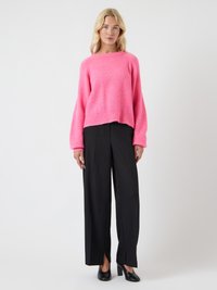 Fényes rózsaszín, oversized pulóver, puha anyagból készült, fekete, széles lábú nadrággal párosítva, amely oldalsó hasítékokkal rendelkezik, és fekete magassarkú cipőkkel.
