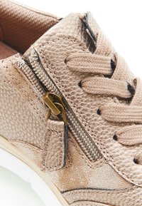 Sneaker metallico oro con texture e zip foderata, lacci marroni. Presenta una suola bianca e dettagli neri vicino ai fori per i lacci.