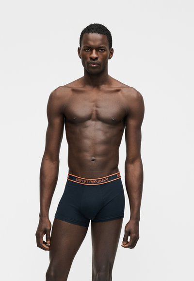 Άνδρας μοντέλο στέκεται μπροστά σε απλό φόντο φορώντας μπλε σκούρα boxer briefs Emporio Armani με πορτοκαλί ζώνη μέσης.