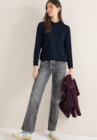 Femme debout tenant une veste violette pliée, portant un sweat à capuche bleu marine, un jean gris délavé et des baskets blanches avec des accents bleus.