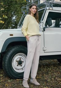 Pull jaune clair texturé, pantalon de jogging gris clair avec poches, et baskets beige, debout à côté d'un véhicule blanc sur un terrain herbeux.