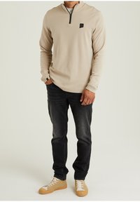 Beiger Pullover mit einem Reißverschluss und einem kleinen schwarzen Logo, kombiniert mit dunklen Jeans und hellbraunen Sneakers. Der Stoff wirkt glatt und hat eine lässige Passform.