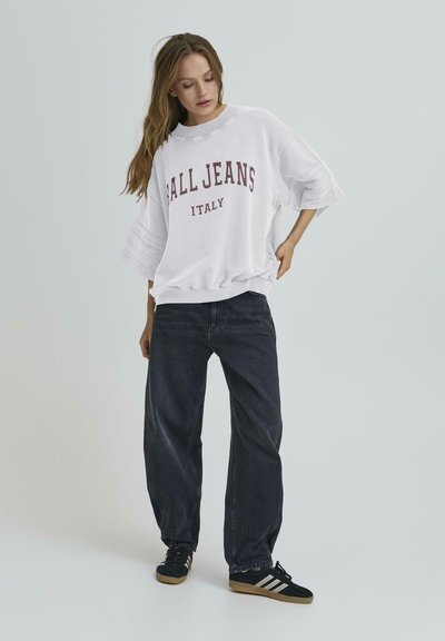 Hanorac alb supradimensionat cu textul "FALL JEANS ITALY" în burgundă, asortat cu blugi largi de culoare albastru închis și adidași negri cu dungi albe.