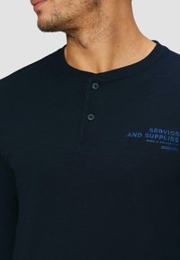 Tumeansininen pitkähihaisessa henley-paidassa, joka on valmistettu pehmeästä kankaasta, on kolme nappia kauluksessa ja rintamuksessa brodeerattu sininen teksti.