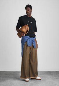 Moški v črnem majici, širokih rjavih hlačah, modrem puloverju, zavezanim okoli pasu, belih čevljih, drži zloženo rjavo usnjeno torbo, stoji na sivi tla.