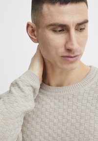 Beige knit sweater with a textured pattern, round neckline, and ribbed cuffs. Model has short hair and is touching their neck.