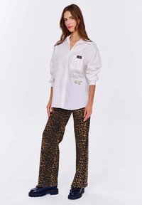 Biała, oversize koszula z guzikami i kieszonką z tekstem. Brązowe, w panterkę, rozszerzane spodnie z teksturowanej tkaniny oraz czarne buty na platformie.