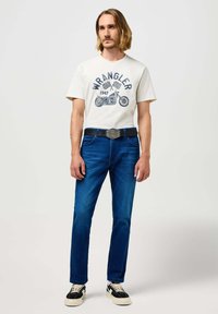 Witte katoenen t-shirt met blauw grafisch ontwerp, gecombineerd met donkerblauwe jeans en zwarte schoenen. Een zwarte riem met zilveren gesp maakt de outfit compleet.