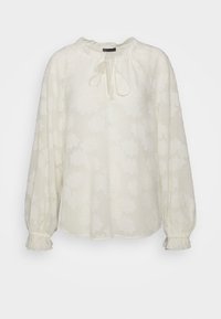 Blouse blanche à manches longues avec motif jacquard floral, col noué et poignets volantés sur fond blanc uni.