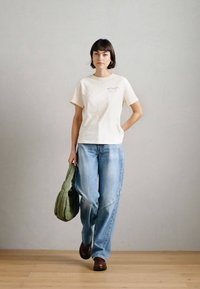 Μπεζ κοντομάνικο μπλουζάκι, μπλε φαρδιά τζιν παντελόνια, καφέ παπούτσια και πράσινη επενδεδυμένη τσάντα. Απλός σχεδιασμός με ελάχιστες λεπτομέρειες.