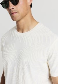 T-shirt bianco a maniche corte con un motivo testurizzato a foglia di palma. Realizzato in morbido tessuto con design a collo rotondo. Messo in risalto da dettagli tonali.