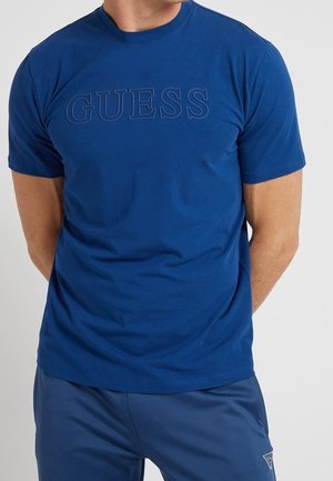 Mężczyzna w niebieskim T-shircie z krótkim rękawem z wypukłym logo "GUESS" oraz pasujących niebieskich spodniach z bocznymi kieszeniami.