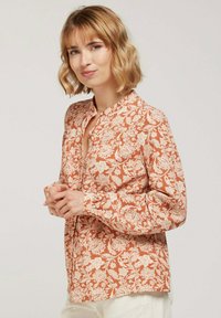 Blouse florale couleur rouille avec imprimé botanique crème, design avec col, fermeture à boutons et manches longues. Tissu doux et coupe décontractée.