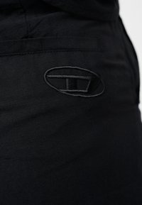 Gros plan sur un tissu noir avec un logo ovale noir brodé contenant une barre horizontale et une ligne verticale.