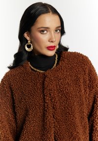 Brun tekstureret jakke med en shaggy finish, båret over en sort turtleneck. Guld kædeting og hoops øreringe fuldender looket.