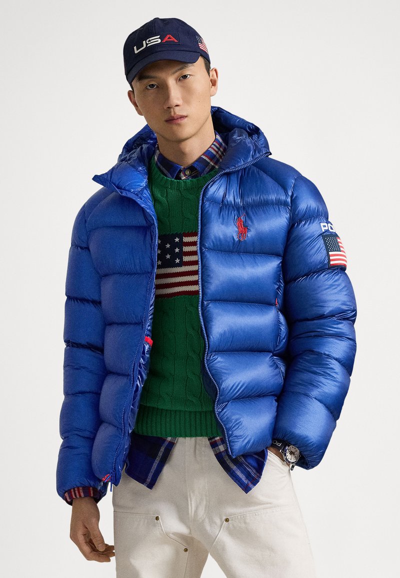 Doudoune bleue avec capuche, présentant une texture brillante, détaillée avec des accents rouges et un écusson du drapeau américain sur la manche.