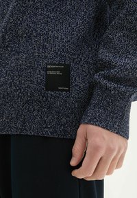 Granatowy sweter z dzianiny z teksturowanym wzorem i ściągaczami. Zawiera czarną metkę z białym napisem na dolnej stronie.