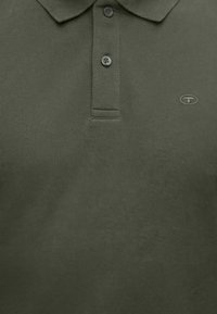 Πόλο μπλούζα σε ελιά πράσινο χρώμα με τρία κουμπιά και μικρό οβάλ λογότυπο στην αριστερή περιοχή του στήθους.