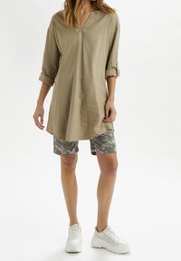 Tan linnen tuniek met een v-hals en opgerolde mouwen, gecombineerd met grijze camouflage shorts en witte sneakers, op een lichte achtergrond.