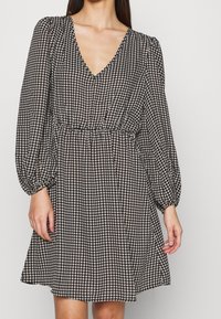 Svart- och vittmönstrad klänning med V-ringning, långa puffärmar och elastisk midja, med en utsvängd kjol och houndstooth-design.